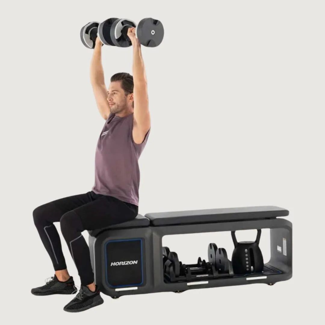 Man performing a seated dumbbell shoulder press on the Horizon fitness bench with gear stored below.