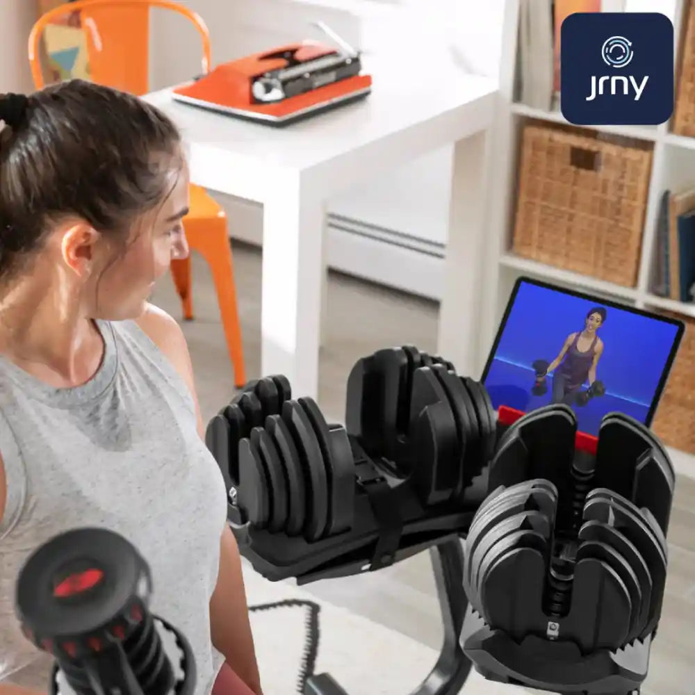Woman using adjustable dumbbells in a home office setting with a laptop showing a fitness video.