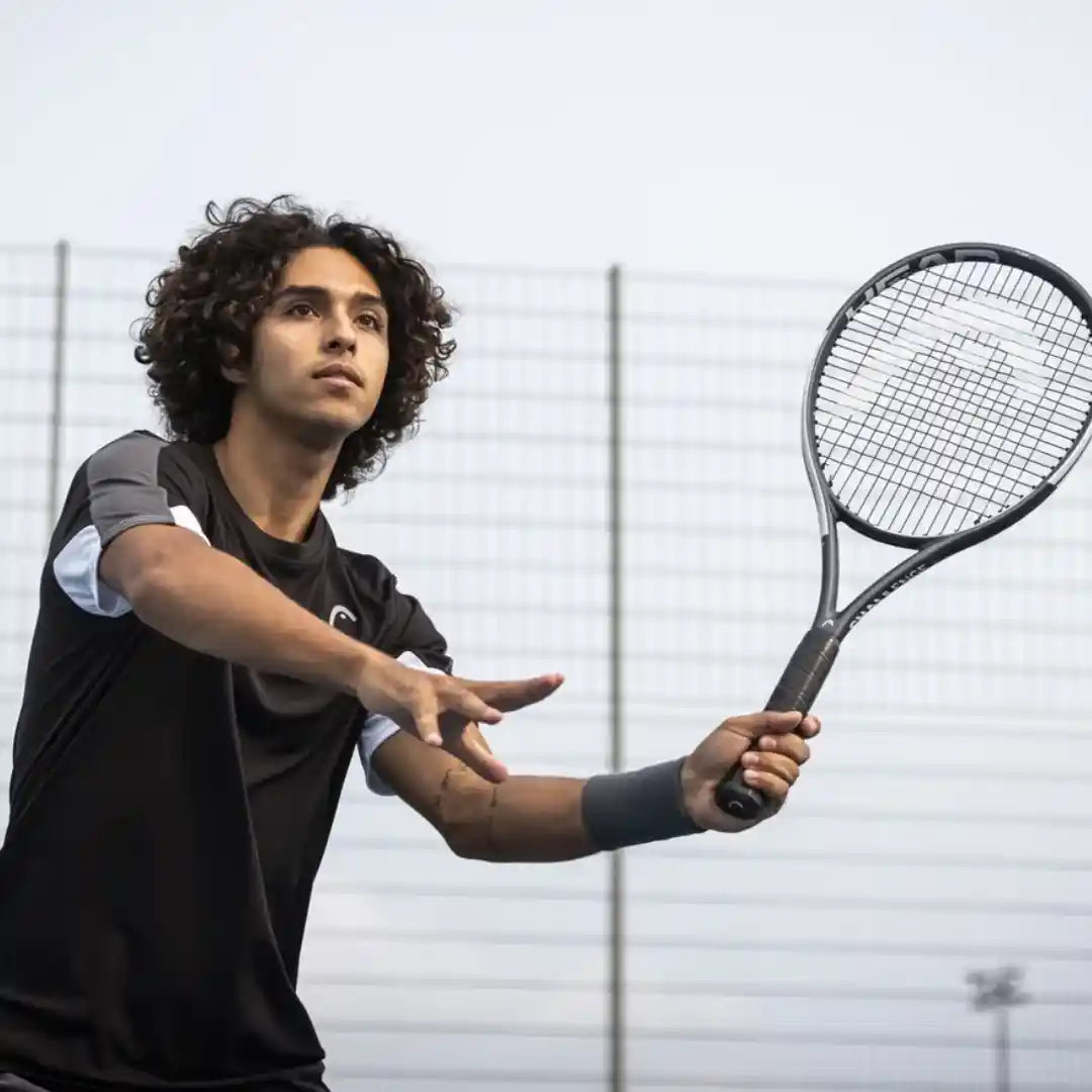 A male tennis player with curly dark hair is captured mid-swing, holding a black tennis racket. He is wearing a black athletic shirt with gray and white accents and a dark gray wristband, set against an outdoor tennis court background with a white fence and a pale sky.